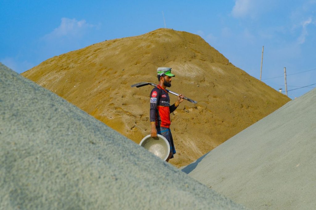 A construction worker carrying tools across sand dunes under a clear, bright sky.