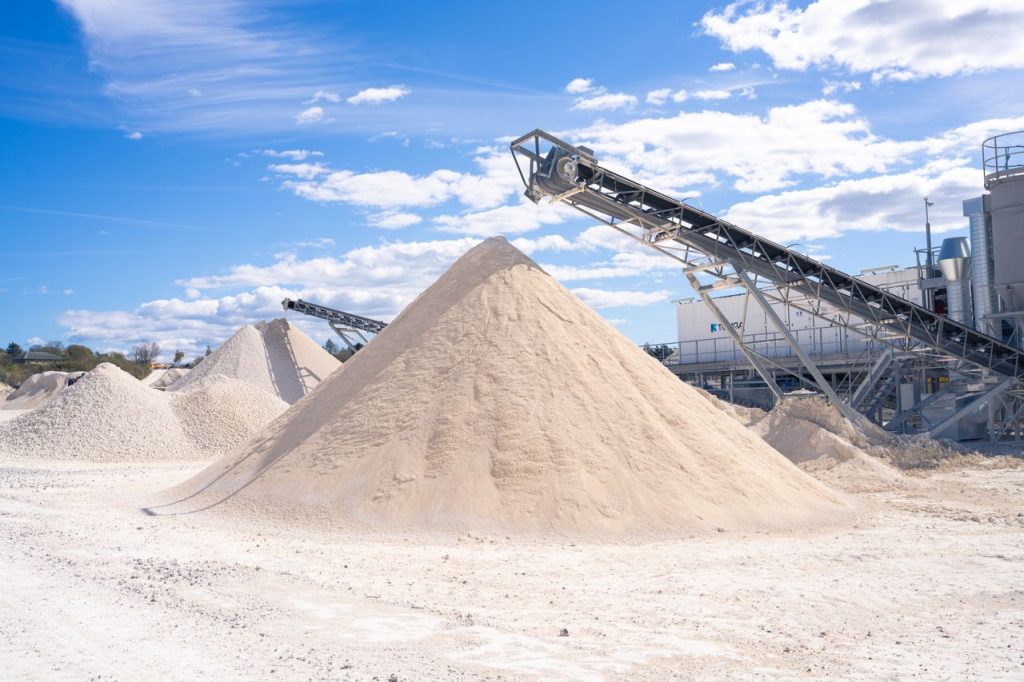 Quarry scene with conveyor belt and large sand piles under a blue sky.
